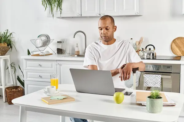 Разнообразный афроамериканец с миастенией и грависом сидит за кухонным столом, поглощённый ноутбуком. — стоковое фото