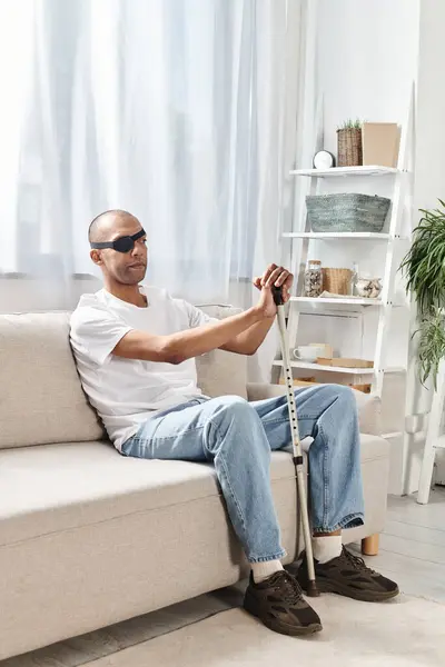 Hombre afroamericano con síndrome de miastenia gravis sentado en un sofá, sosteniendo un bastón, profundamente en el pensamiento. - foto de stock