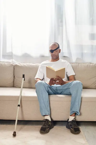 Un hombre con Miastenia Gravis, una afroamericana, está absorto en la lectura de un libro mientras está sentado cómodamente en un sofá. - foto de stock