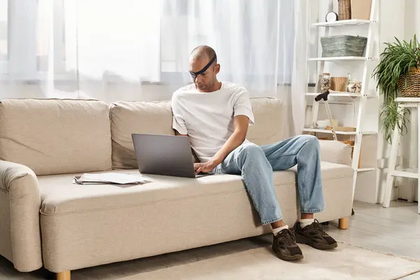 A man with myasthenia gravis syndrome sits on a couch, engrossed in his laptop. — Stock Photo