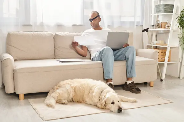 Um homem, lutando contra a miastenia gravis, senta-se em um sofá com um laptop, acompanhado por seu cão Labrador leal.. — Fotografia de Stock