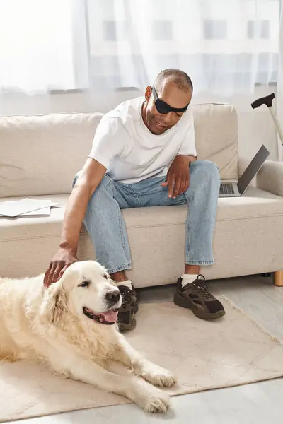 A disabled African-American man with myasthenia gravis syndrome sits next to a loyal Labrador dog on a couch. — Stock Photo