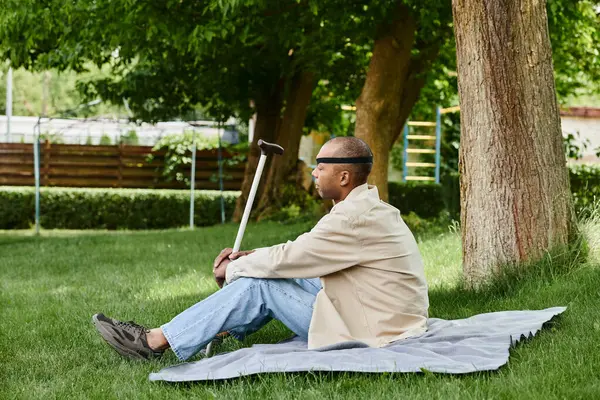 Инвалид афроамериканец с синдромом миастении и грависа сидит на покрывале в траве, принимая разнообразие и включение. — стоковое фото