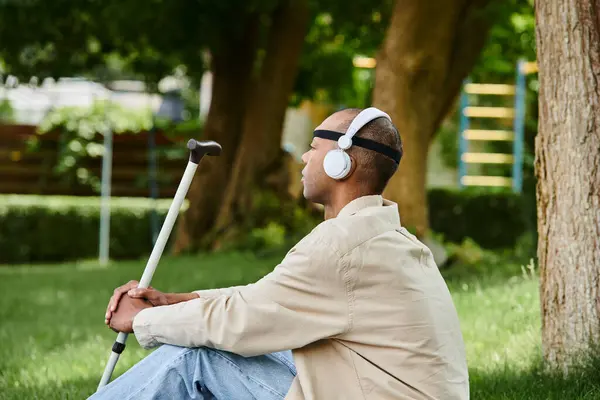 Um homem afro-americano com síndrome da miastenia gravis senta-se na grama com fones de ouvido, curtindo música. — Fotografia de Stock
