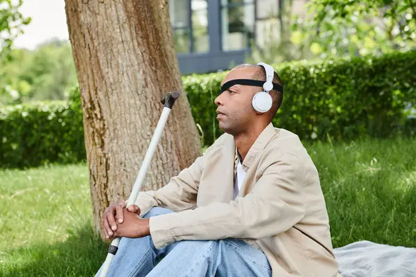 Un hombre con auriculares está sentado en una manta junto a un árbol, disfrutando de la música y el entorno tranquilo - foto de stock