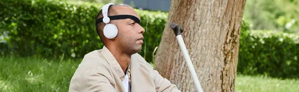 Un hombre con auriculares disfruta de la música junto a un árbol, mostrando diversidad e inclusión. - foto de stock