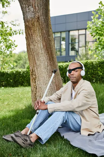 Un hombre con miastenia gravis se sienta debajo de un árbol, escuchando música con auriculares - foto de stock