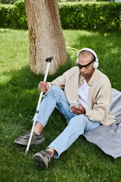 Un hombre afroamericano con síndrome de miastenia gravis se sienta en la hierba, escuchando música a través de auriculares. - foto de stock