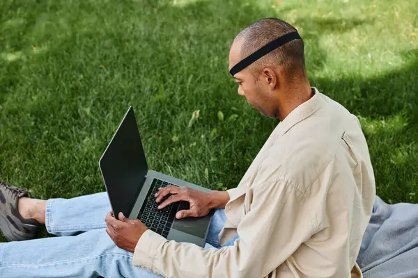 Инвалид афроамериканец с синдромом миастении, работающий на ноутбуке, сидя на зеленой траве — стоковое фото