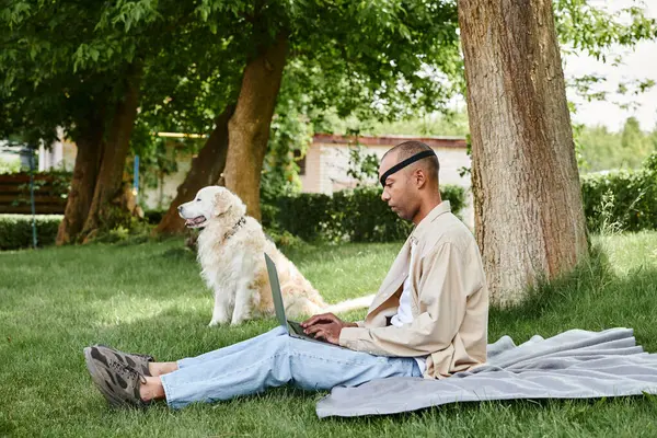 Un homme diversifié atteint de myasthénie Gravis est assis sur l'herbe, à l'aide d'un ordinateur portable accompagné de son fidèle chien Labrador. — Photo de stock