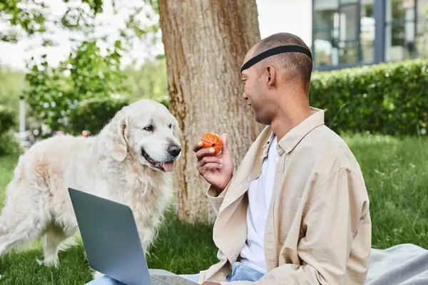 Un uomo afroamericano con la sindrome della miastenia grave, che lavora su un computer portatile nell'erba con il suo fedele cane Labrador al suo fianco. — Foto stock