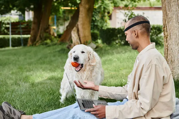 Un hombre afroamericano con miastenia gravis se sienta en la hierba con un portátil, balanceando una pelota en su boca mientras su Labrador mira. - foto de stock