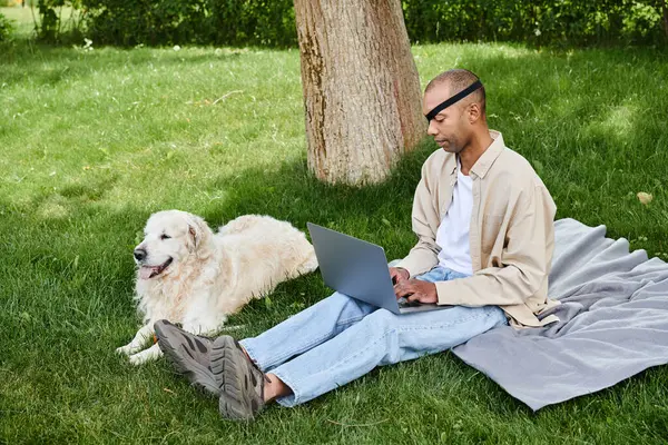 A man with Myasthenia Gravis syndrome sits in the grass, working on a laptop beside his loyal Labrador dog. — Stock Photo