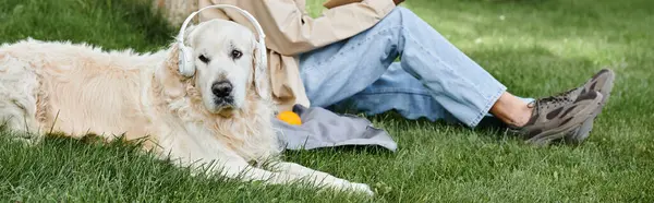 A disabled African American man sitting in the grass with his loyal Labrador dog in headphones by his side. — Stock Photo