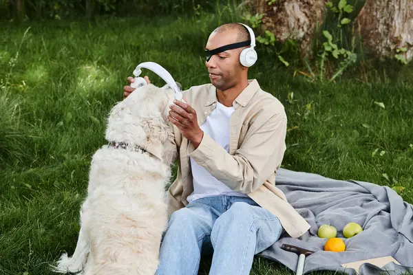 African American man with myasthenia gravis sitting on a blanket, enjoying music with Labrador dog wearing headphones. — Stock Photo