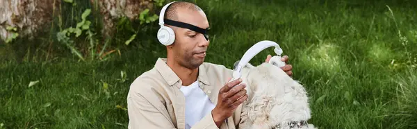 An African American man with myasthenia gravis syndrome relaxes in the grass with a Labrador dog wearing headphones. — Stock Photo