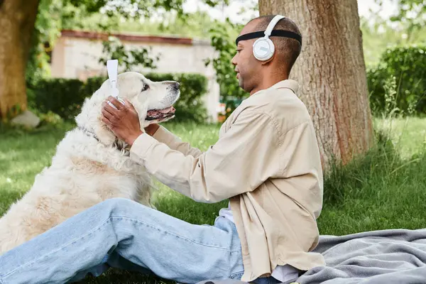 Un Afro-Américain handicapé atteint du syndrome de la myasthénie grave est assis dans l'herbe avec un chien Labrador portant un casque, profitant d'un moment paisible ensemble. — Photo de stock
