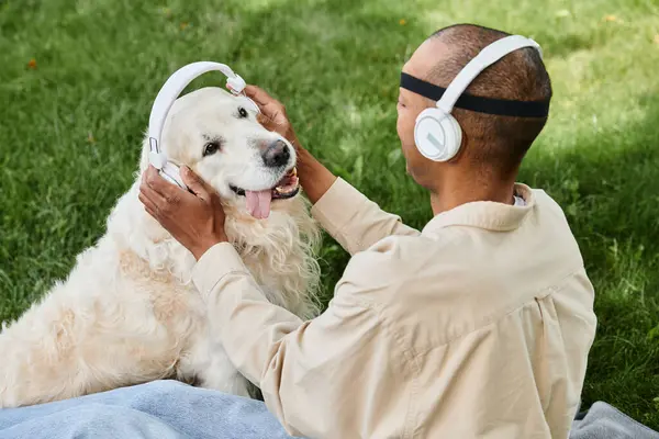Разнообразный человек с синдромом миастении и грависа сидит на траве и радостно гладит свою верную собаку-лабрадора, в то время как оба носят наушники. — стоковое фото