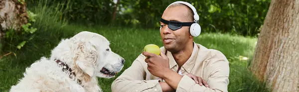 Un afroamericano discapacitado con síndrome de miastenia gravis escucha los auriculares mientras come una manzana junto a su leal perro Labrador. - foto de stock