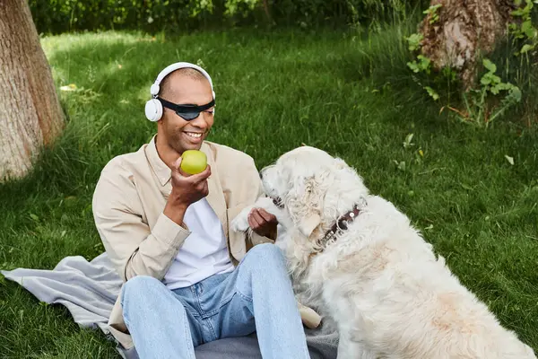 A disabled African American man with myasthenia gravis syndrome sits on a blanket with a Labrador dog, enjoying an apple. — Stock Photo