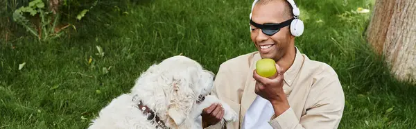 Un hombre afroamericano diverso con miastenia gravis sostiene una manzana mientras su perro Labrador está junto a él. - foto de stock