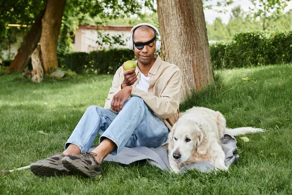 Un hombre afroamericano con síndrome de Miastenia Gravis se sienta en la hierba junto a un perro labrador y una manzana. - foto de stock