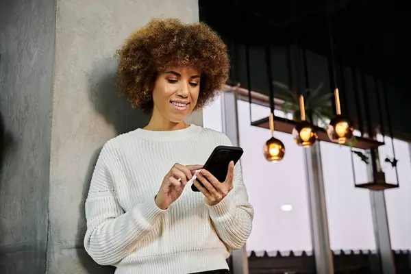 Афроамериканка в белом свитере, завораживающая своим мобильным телефоном в современном кафе. — стоковое фото