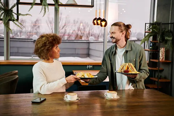 Афроамериканка и мужчина садятся за стол с едой в современном кафе, беседуют и наслаждаются обедом вместе. — стоковое фото
