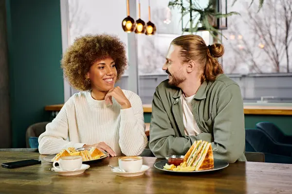 Афроамериканка и мужчина сидят за столом, набитым тарелками еды, в современном кафе, наслаждаясь совместным обедом. — стоковое фото