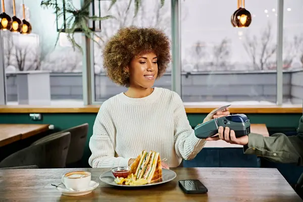 Una mujer afroamericana está sentada en una mesa, delicadamente preparada con un plato de deliciosa comida frente a ella. - foto de stock