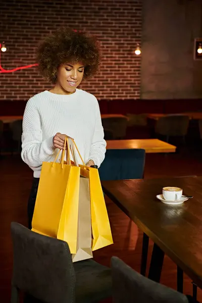 A stylish African American woman holding a yellow shopping bag in a trendy restaurant, surrounded by modern decor — Stock Photo