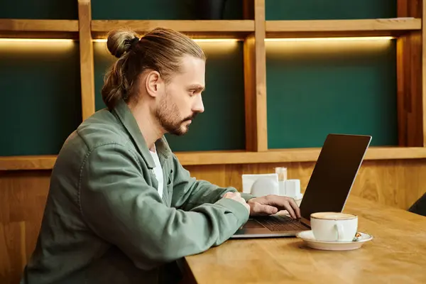 Un hombre se sienta en una mesa en un café moderno, absorto en su trabajo en una computadora portátil. - foto de stock