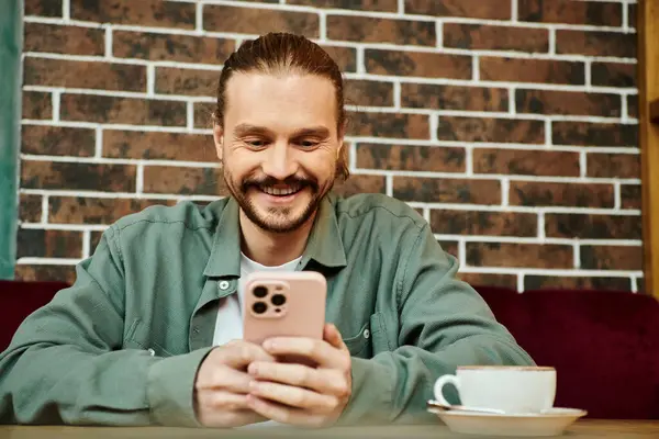 Hombre feliz está sentado en una mesa en un café moderno, centrado en el uso de su teléfono celular. - foto de stock