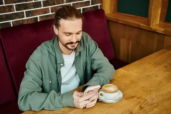 Un hombre se sienta en una mesa, saboreando una taza de café en un café moderno y usando un teléfono inteligente - foto de stock