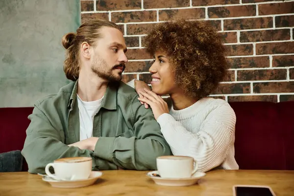 Una mujer afroamericana y un hombre sentado en una mesa, disfrutando de tazas de café en un moderno café. - foto de stock