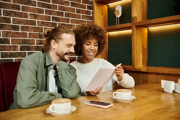 Una donna afroamericana e un uomo si siedono a un tavolo, concentrati su un menu in un ambiente moderno caffè. — Foto stock