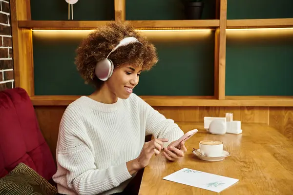 An African American woman with headphones sits at a table in a modern cafe, lost in her music. — Stock Photo