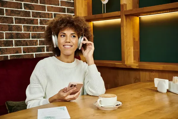 Una mujer afroamericana se sienta en una mesa de café, hablando por su teléfono celular, conversando. - foto de stock