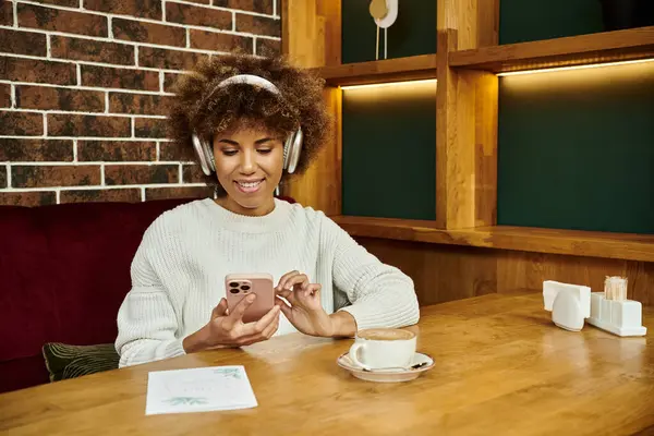 Una mujer afroamericana se sienta en una mesa en un café moderno, usando auriculares y sumergiéndose en su propio pequeño mundo. - foto de stock