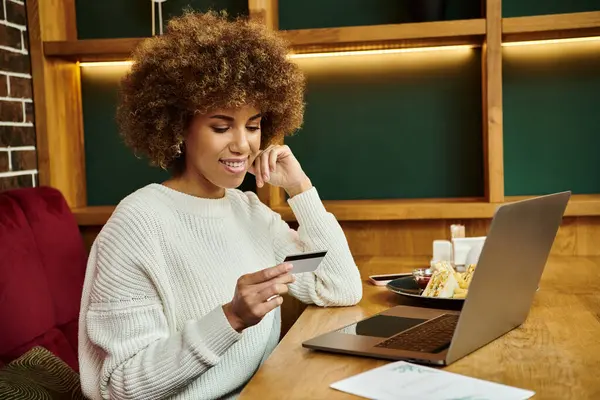 Una moderna donna afroamericana si siede in un caffè, concentrandosi intensamente sulla sua carta di credito vicino al computer portatile — Foto stock