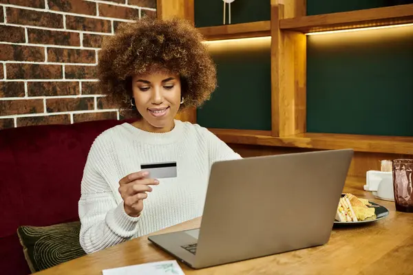 Una mujer afroamericana moderna sentada en una mesa con un portátil y una tarjeta de crédito, haciendo compras en línea. - foto de stock