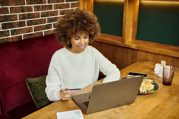 Una mujer afroamericana se sienta en una mesa en un café moderno, enfocada en la pantalla de su computadora portátil. - foto de stock