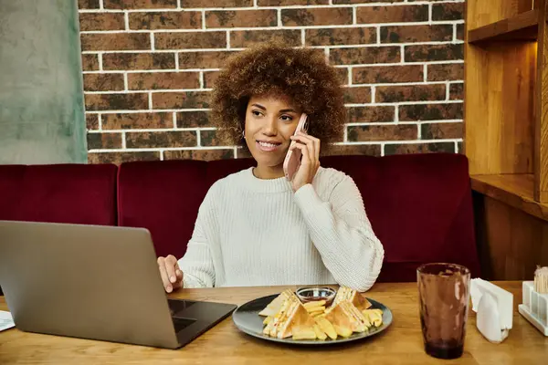 Una donna afroamericana si siede a un tavolo con un computer portatile e un piatto di cibo, focalizzato sul suo lavoro. — Foto stock