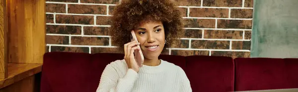Una donna afroamericana elegante si siede su un moderno divano da caffè, impegnata in una conversazione telefonica. — Foto stock