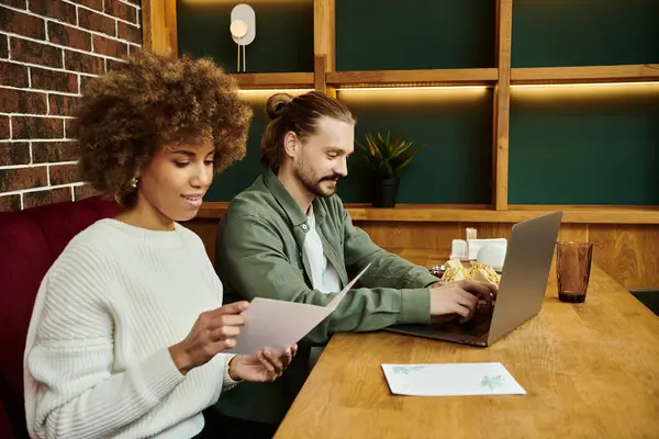An African American woman and a man sit at a table with a laptop, engaged in conversation and collaboration in a modern cafe setting. — Stock Photo