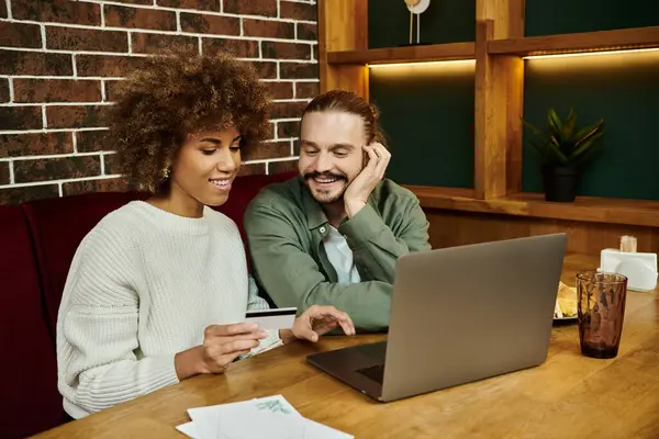 Une femme afro-américaine et un homme sont assis à une table dans un café moderne, absorbé par la recherche d'un écran d'ordinateur portable. — Photo de stock