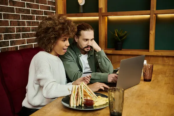 A man and African American woman sit at a table, engrossed in the content on a laptop screen in a trendy cafe. — Stock Photo