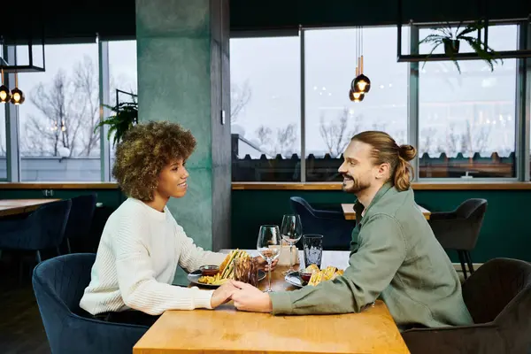 Una mujer afroamericana y un hombre disfrutan de una comida juntos en un café moderno, comprometidos en la conversación y la risa. - foto de stock