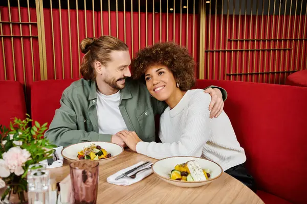 Una mujer afroamericana y un hombre disfrutan de una comida juntos en una mesa llena de deliciosa comida en un moderno entorno de cafetería. - foto de stock