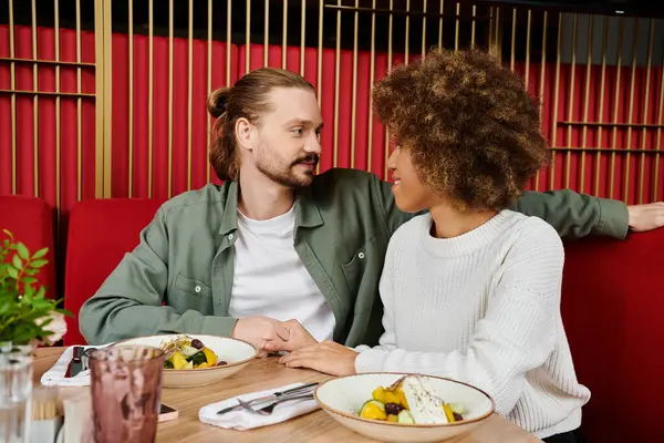 Una donna afroamericana e un uomo si siedono a un tavolo con piatti di cibo in un moderno caffè. — Foto stock